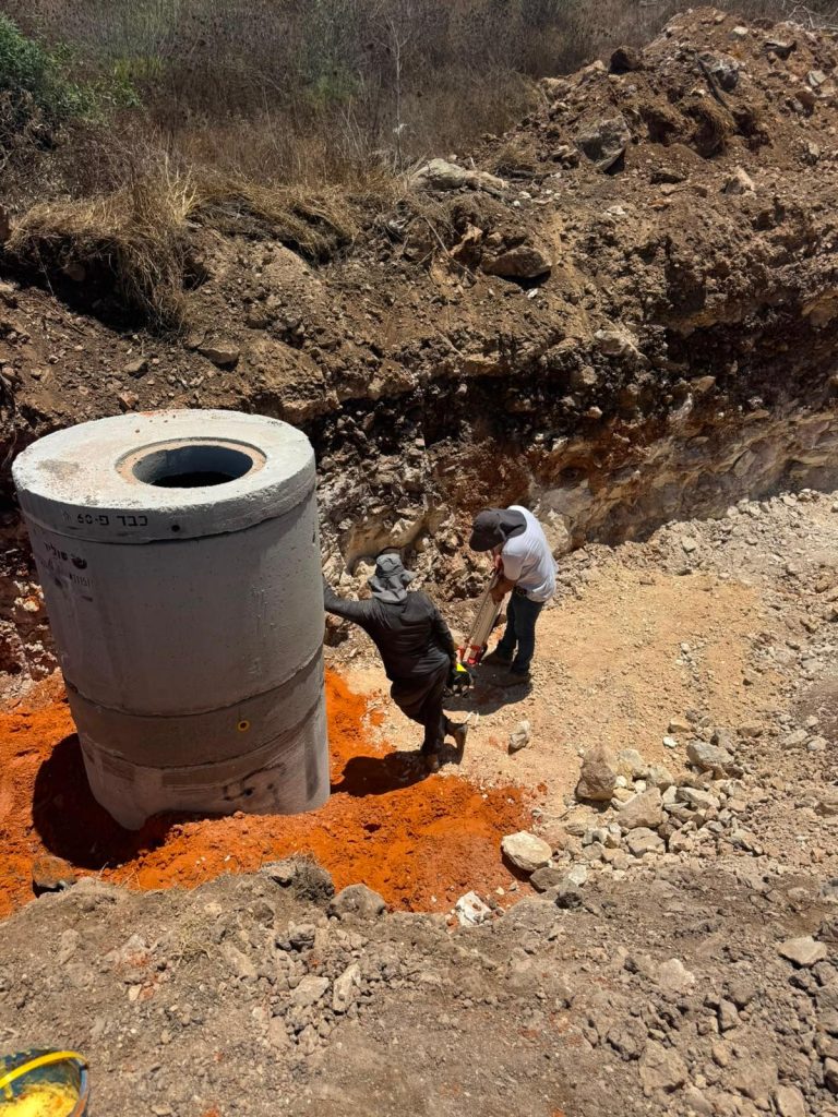 שני עובדים עם אלמנט בטון טרומי של השוחה המותקן בתוך החפירה, כשסביבו בוצע מצע חול לייצוב והונחה רצועת סימון תת-קרקעית לאורך תוואי הקו.